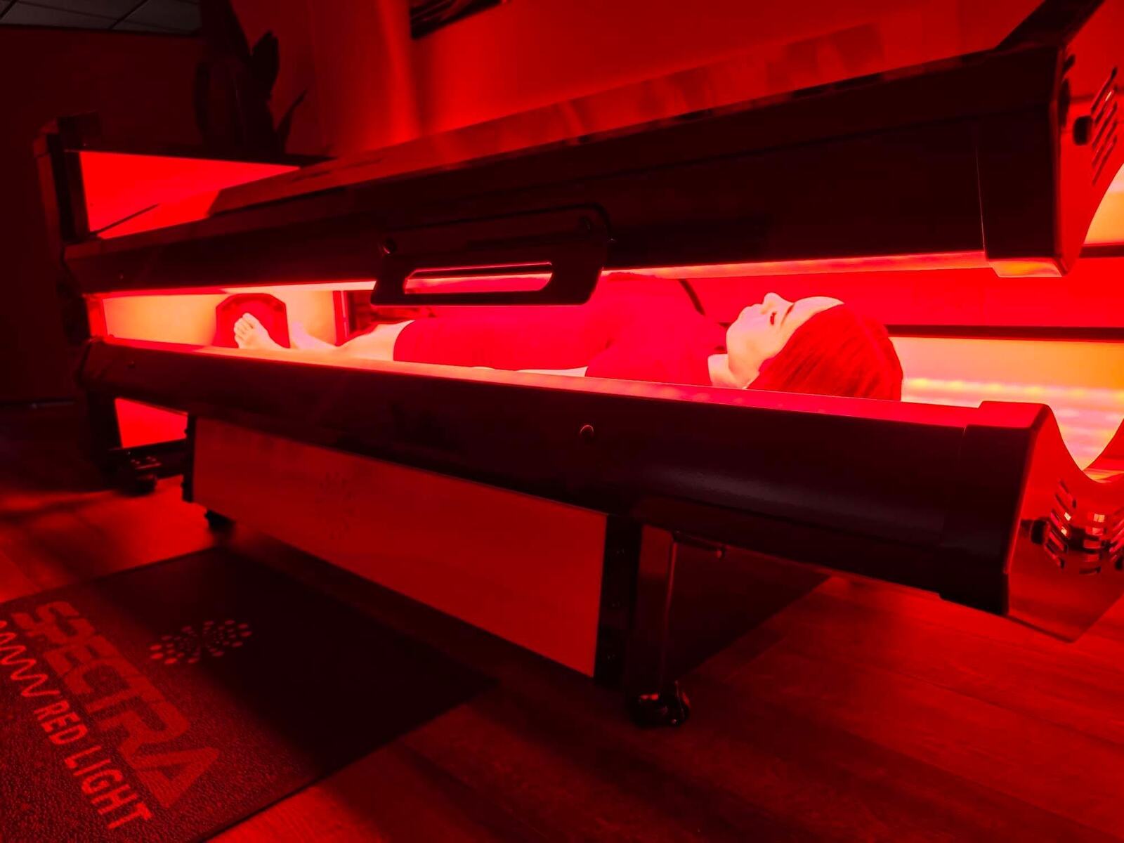 Side view of a guest in the Spectra S10 Pro red light bed, lit in deep red, with a Spectra Red Light mat on the floor